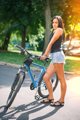 Naklejka premium Young pretty brunette girl in denim shorts with a bicycle on the street in summer.