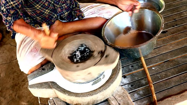 upper view on woman sat cross-legged using hand-turned millstone to grind wet rice for making soaked rice flour