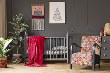Patterned armchair next to baby's bed with red blanket in grey bedroom interior with plants. Real...
