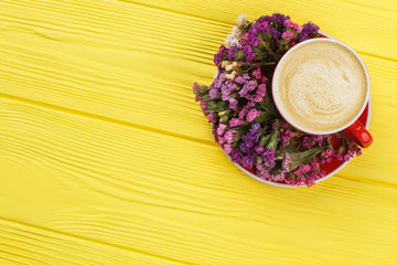 Obraz na płótnie Canvas Mug of coffee and limonium statice flowers. Top view, flat lay. Yellow wooden background.