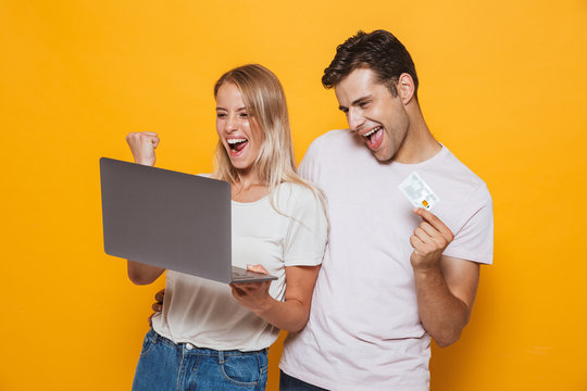 Young loving couple isolated over yellow wall background using laptop computer make winner gesture holding credit card.