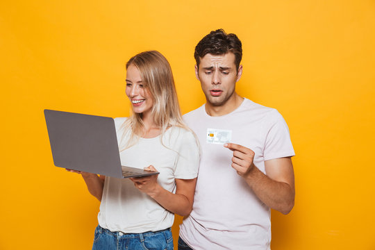 Woman isolated over yellow wall background using laptop computer while her confused sad boyfriend holding credit card.
