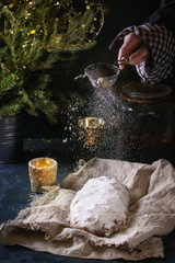 Whole Christmas cake, German festive baking. Stollen on linen napkin with sprinkling sugar powder by vintage sieve in child hand on dark blue table. Fir tree and candles at background. Rustic style