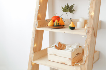 Orange pumpkins on plate, dry autumn leaves in glass vase, candles, wooden box with sweater and glowing garland on wooden rack. Light stylish cozy fall home interior decor. Autumn decoration.