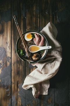 Traditional Japanese Noodle Soup with shiitake mushroom, egg, sliced beef and greens served in ceramic bowl with wooden chopsticks and white spoon on cloth. Dark wooden background. Flat lay, space.