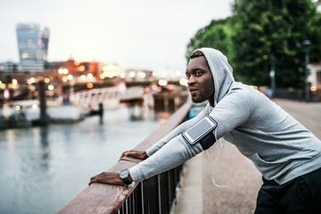 Black man runner with smartphone in an armband on the bridge in a city, resting.