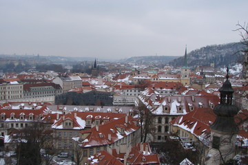 Fototapeta na wymiar The landscape from the height of the mountain on the snow-capped houses, churches and castles of the historic part of the city of Prague, surrounded by hills covered with forest.
