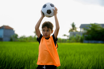 lifestyle portrait of handsome and happy young boy holding soccer ball playing football outdoors at green grass field smiling cheerful wearing training vest