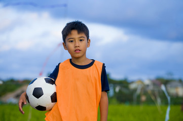 lifestyle portrait of handsome and happy young boy holding soccer ball playing football outdoors at green grass field smiling cheerful wearing training vest