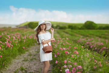 Beautiful young woman in white hat with long curly hair holding basket posing over spring blossom pink roses garden. Attractive charming brunette female outdoor portrait.