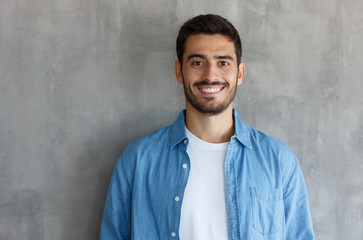Horizontal shot of handsome European guy pictured isolated on gray background standing against wall in blue denim shirt, looking straight at camera, feeling very confident and relaxed, smiling - Powered by Adobe