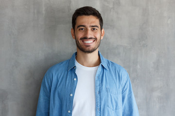 Horizontal closeup of young handsome European Caucasian male isolated on grey background wearing blue denim shirt and white T-shirt, having short and face hair, smiling confidently and friendly