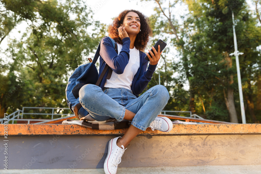 Poster Woman sitting outdoors in park using mobile phone listening music with earphones.