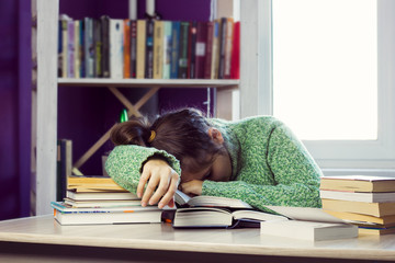 woman in a green sweater sleeps at a table in the middle of a stack of books and study materials, a blurred bookcase and a window for the background