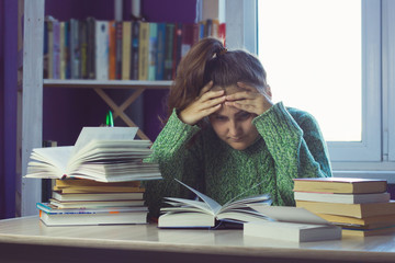 sad woman in a green sweater at a table in the middle of a stack of books and study materials, a blurred bookcase and a window for the background - obrazy, fototapety, plakaty