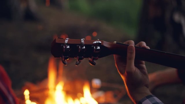 Close-up shot of male tourist's hand playing the guitar during evening at campsite with fire burning in background. Musical instruments, nature and people concept.