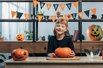 portrait of smiling kid at tabletop with pumpkins and hanging happy halloween flags behind at home