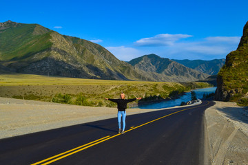 Young pretty blonde woman on a black asphalt road with yellow markings and sand on the roadside with his arms outstretched behind a picturesque landscape of Altai moumtains and turquoise river Katun