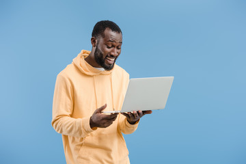 happy young african american man with closed eyes holding laptop isolated on blue background