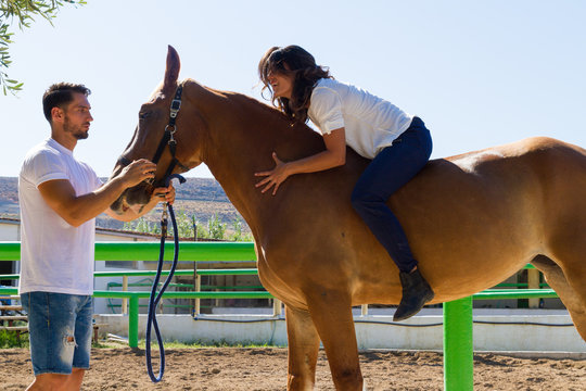 Young woman takes horse riding lessons from young male instructor in the riding club. Young couple with brown horse at the stables. Sports Liferstyle concept.