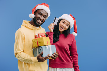 cheerful african american man in christmas hat holding gift boxes while his girlfriend showing credit card isolated on blue background