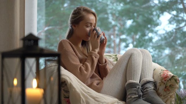 Cozy winter lifestyle. Happy young woman enjoy of cup of hot coffee sitting home by the big window with winter snow tree background