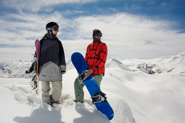 guy and a girl in warm clothes stand with snowboards against the mountain snow tops
