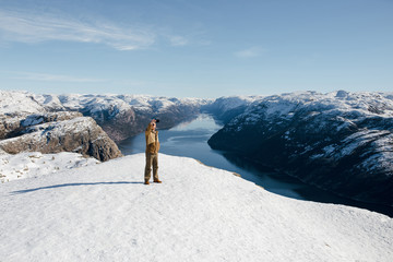 Traveller man standing on the Preikestolen cliff edge and making selfies with the smartphone of the beautiful landscape view of mountains and fjord on background. Norway
