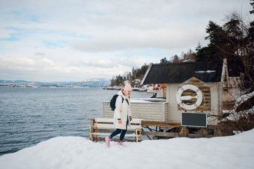 Beautiful young blonde woman in white winter jacket and pink hat walking near the traditional Norwegian house on the sea shore on winter day. Fjord.