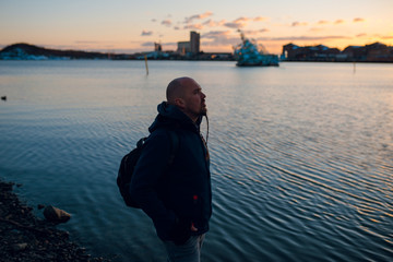 Traveler man with backpack standing on the sea coast and looking at the bay at sunset. Dusk. Cold winter day in Norway, Oslo