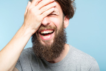 facepalm. happy smiling joyful man covering his face. shame and fun concept. portrait of a young bearded guy on blue background. emotion facial expression.