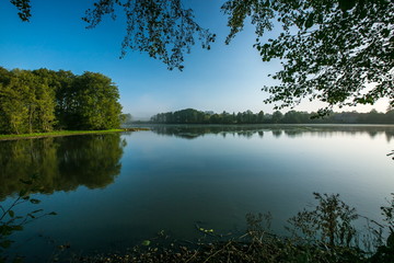 Bright colorful landscape with lake and blue water with reflection of green trees, sunny day, clear sky, framed with branches, horizon in distance with trees, calm water surface