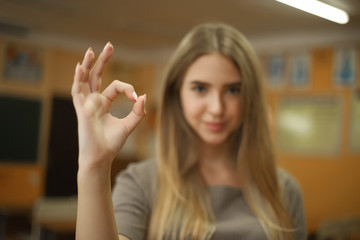 Student schoolgirl lady with long hair standing in empty classroom looking camera showing okay gesture. Focus on hands.