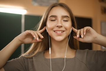 Smiling young student schoolgirl lady with long hair standing in empty classroom listening music with earphones.