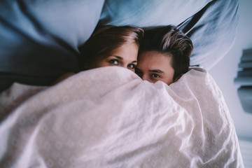 young couple hiding under blanket in bed
