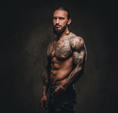 Portrait of a shirtless bearded tattooed male posing with hand in pocket. Isolated on dark textured background.