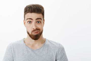Portrait of silly and funny handsome man with beard, moustache and blue eyes smirking making uncertain awkward face looking in mirror and thinking about making changes posing against gray background