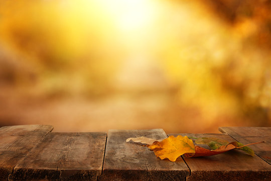 image of front rustic wood table with dry gold leaves and fall bokeh background.