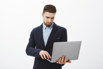 Focusing on business. Portrait of smart and ambitious concentrated good-looking young male entrepreneur with beard and blue eyes holding laptop in hand browsing checking schedule with determined look