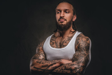 Brutal bearded tattooed male in white shirt posing with crossed arms. Isolated on a dark textured background.