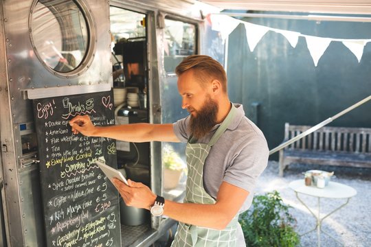 Male waitress writing menu on menu board while using digital