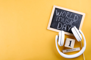 White headphones & blackboard with world music day text. Simple composition w/ headset, chalkboard & wooden calendar showing October first on yellow background. Close up top view, copy space, flat lay