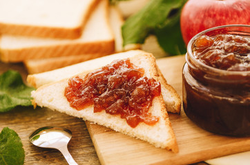 Apple jam on toast and in jar, fresh red apples on a cutting board on a wooden table. Delicious breakfast, rustic style.