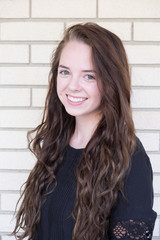 Cute brunette with long hair and black dress looking at camera and smiling, shoulders turned. White brick wall background. Portrait of cute Caucasian girl with long curly hair, head shot of a model.