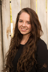 Fototapeta premium Brunette woman posing in front of a rustic old wooden fence wearing a black dress standing. Portrait of a young white woman standing in front of a fence outside in the summer, natural lighting.
