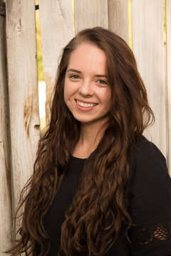 Brunette woman posing in front of a rustic old wooden fence wearing a black dress standing. Portrait of a young white woman standing in front of a fence outside in the summer, natural lighting.
