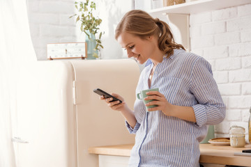 Fototapeta na wymiar Young woman using smartphone leaning at kitchen table with coffee mug and organizer in a modern home. Smiling woman reading phone message. Brunette happy girl typing a text message.