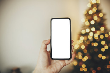 hand holding phone with empty screen on background of golden beautiful christmas tree with lights in festive room. christmas mockup with space for text. advertising, app template