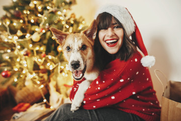 happy girl in santa hat hugging with cute dog on background of golden beautiful christmas tree with lights in festive room. family warm atmospheric moments. winter  holidays