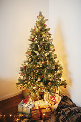 stylish christmas gift boxes under beautiful christmas tree with ornaments, golden lights and presents in festive room. red and gold balls on pine tree branches. decor for winter holidays